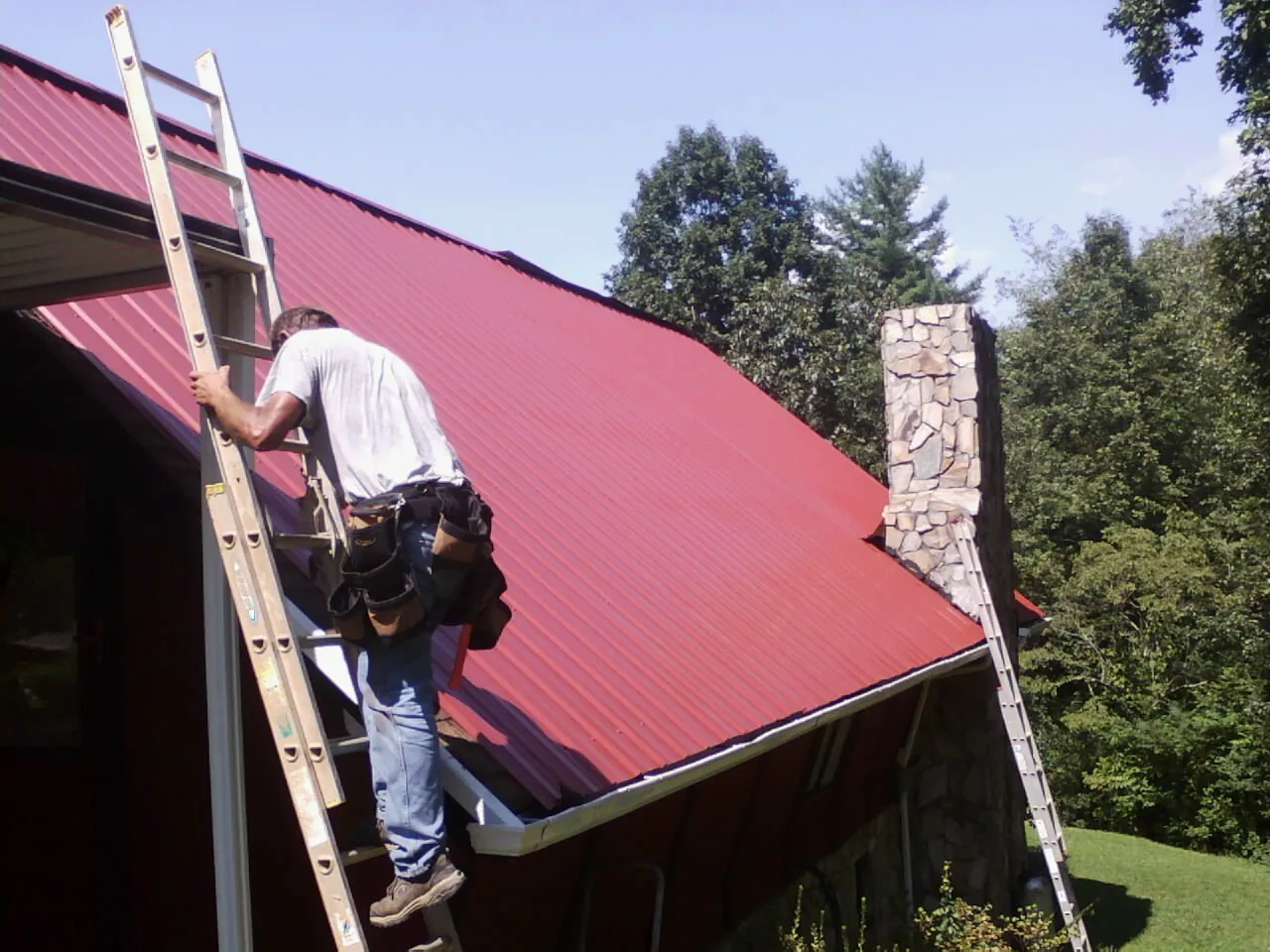 Roofer on a ladder beside a red metal roof and stone chimney — Soffit & Fascia Repair by Roofing Brothers in Westfall